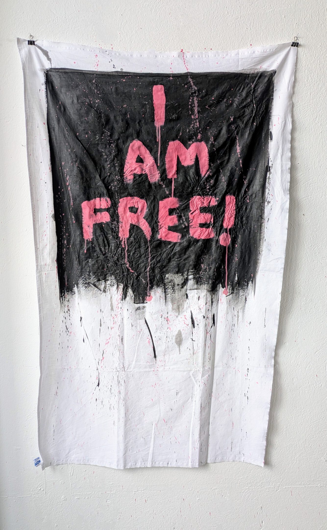 Laura Müller_I am Free_83 x 136 cm_Acrylics on GDR tablecloth_2025 Kunstwerk von Laura Müller "I am free". Der Titel des Bildes steht in Rosa auf Schwarzem Grund. Beides ist auf ein weißes Tischtusch gemalt